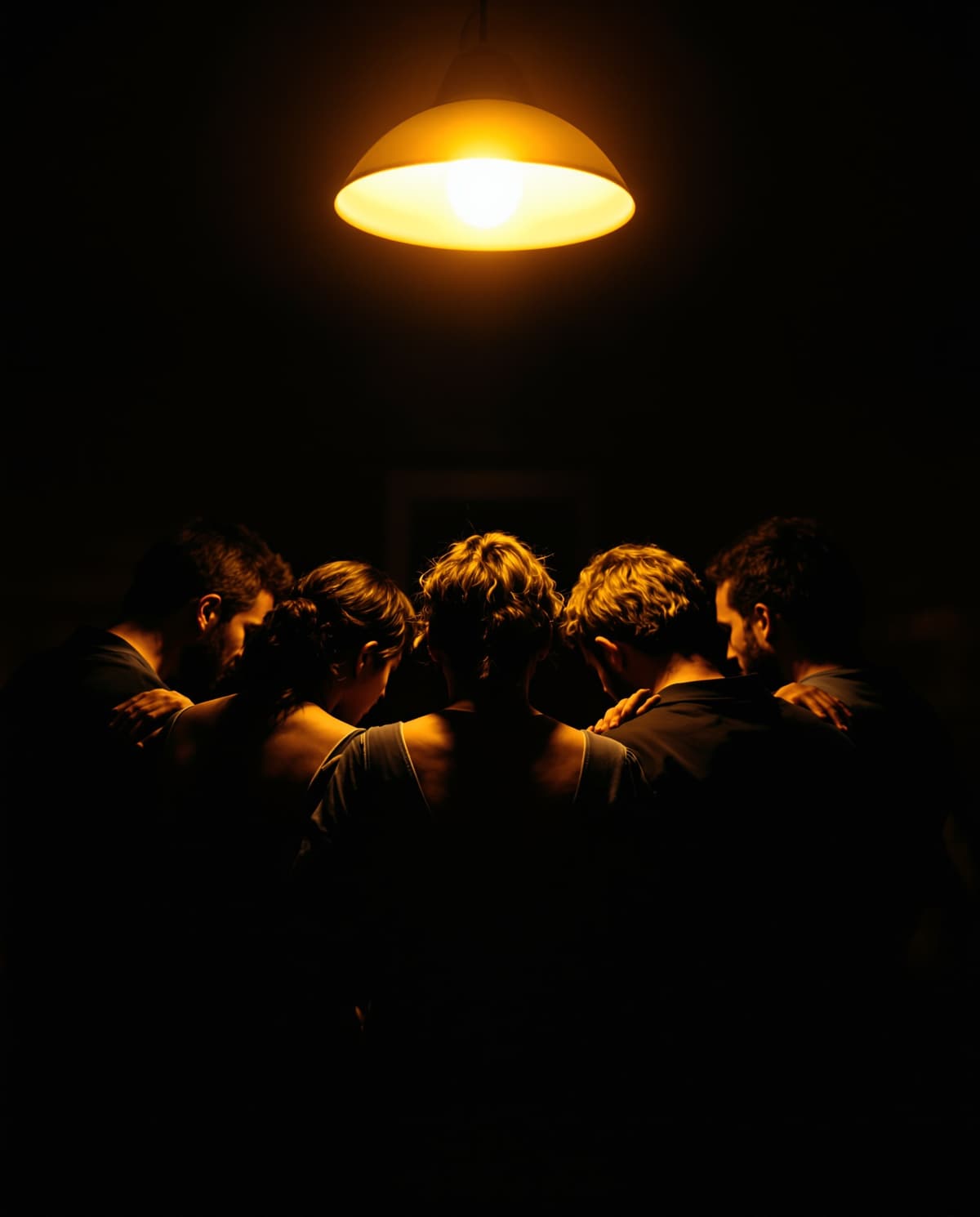 A circle of people with their arms around each other, heads bowed, under a single warm pendant lamp